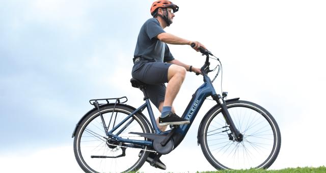 A man wearing a bright orange helmet rides an e-bike on a green field against a blue-sky backdrop. He is wearing a casual t-shirt, trainers and shorts. 