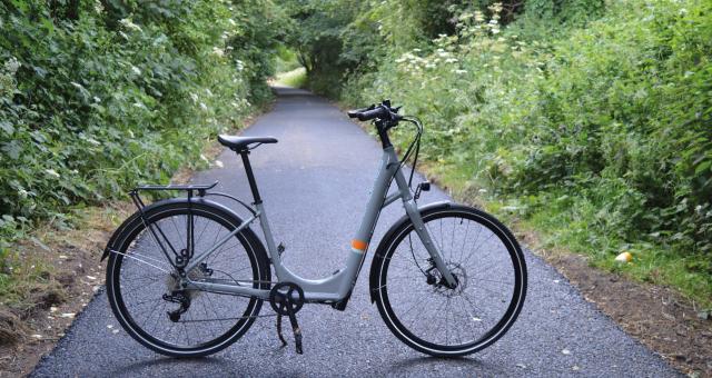 A grey step-through e-cycle stands in the middle of a cycle trail between a wooded area