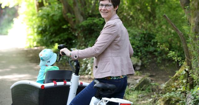 Saskia smiles at the camera as she stands with her cargo bike, transporting her children. She is wearing glasses, a beige jacket and jeans. 