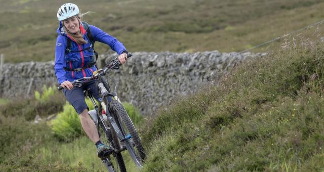 Sophie mountain biking on the Great North Trail. She is wearing a white helmet, blue jacket and grey shorts. 