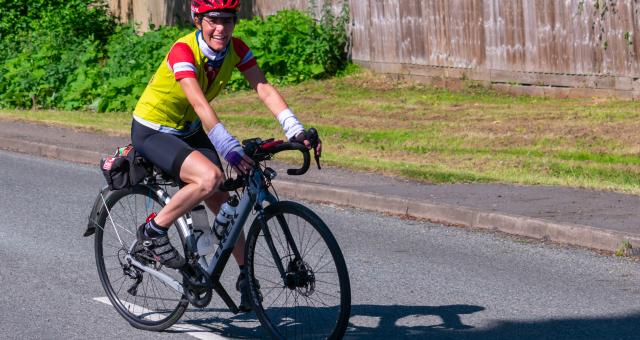 Yasmin smiling as she rides a white Trek bike on road. She is wearing a red helmet, yellow gilet over a red jersey and black cycling shorts. 