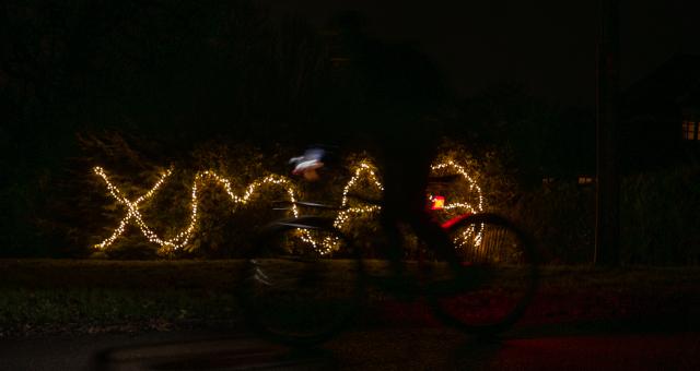 The word 'XMAS' is written out in fairy lights in the background. In front, a cyclist rides past in a blur