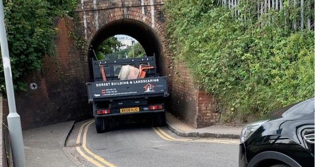 A flat-bed truck is trying to squeeze through a very narrow railway bridge