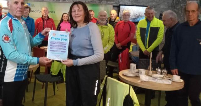 A man and woman pose for the camera holding a certificate of thanks, a group of cyclists stand behind in a cafe