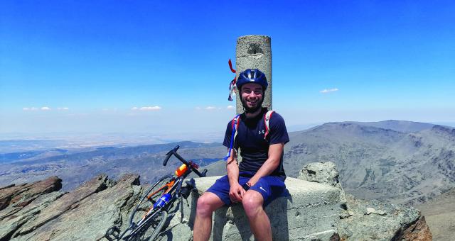 A young man sits at the summit of a mountain with his bicycle next to him. 