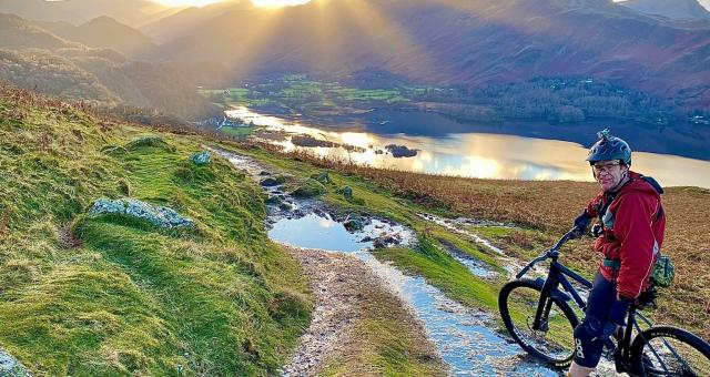 Man stands astride a mountain bike in front of a spectacular view of the sun setting behind a mountain and a trail heading down towards a lake