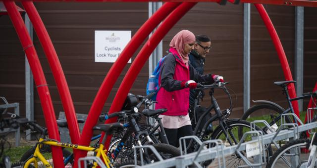 A woman pushes her bike into a bike rack in a red bike shelter. Man in background doing the same. 