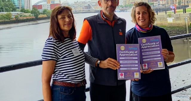 Three people standing side-by-side at Newcastle riverside. Two of them are holding certificates