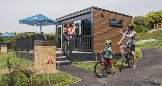 A family waving at each other, next to a glamping pod and on bikes. 