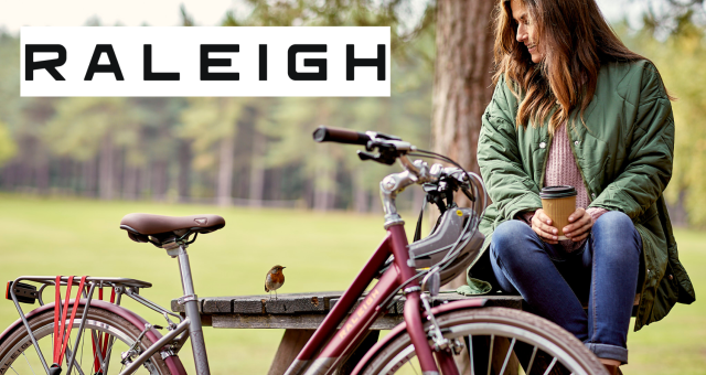 Woman sitting on a bench holding a coffee looking at a robin while her bike leans against the bench