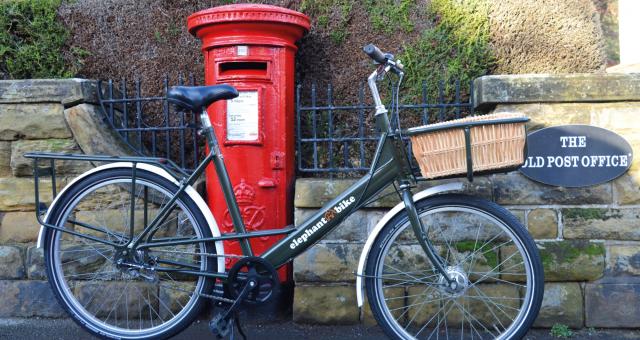 A dark green Elephant Bike. It's an upright, step-through bike with mudguards, long rear rack and a parcel rack with wicker basket at the front