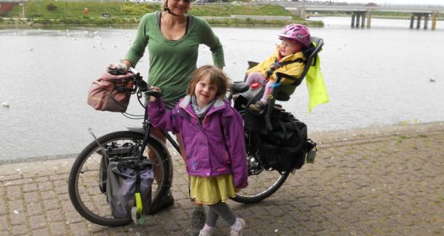 A woman in a green top is standing holding a bike. She has two little girls with her. One is standing in front of the bike and is wearing a purple coat and yellow dress, the other is in a child seat on the back of the bike