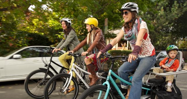 Three women are cycling along an urban road on e-cargo bikes. One has a child on the back of the bike. They are all wearing normal clothes and cycling helmets. There's a white car and trees in the background