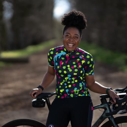 A woman is standing with her road bike. She is perched on the crossbar. She is wearing black cycling shorts and a black and multicoloured polka dot cycling jersey. She is smiling.