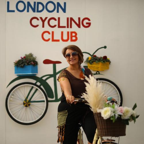 A woman is on a cream Dutch-style bike. The bike’s front basket is full of flowers. She is wearing leggings with a scarf tied around her waist and a brown T-shirt. She has on sunglasses and is smiling at the camera