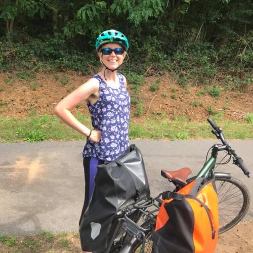 A woman is standing next to a touring bike. She is wearing a blue strappy top with white flower pattern and blue and black leggings, a turquoise cycling helmet and sunglasses. The bike is green and it has two big panniers on the rear rack