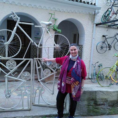 A woman is standing at the gates of a hotel in Spain. The building is covered with old bicycles and even the gates are made from bike frames. She is wearing black trousers, a bright cerise cardigan and blue top, as well as a brightly patterned scarf