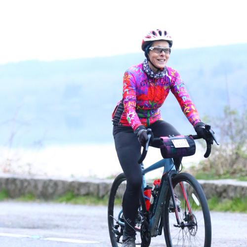 A woman is cycling along the road. She is on a road bike which has a number attached to a handlebar bag; she is taking part in an organised cycling event. She is wearing black cycling leggings and a very bright multicoloured jersey