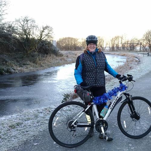 A woman is standing with a light grey Trek mountain bike. It’s winter. She is wearing a warm winter jacket and trousers. Her bike is decorated with blue tinsel. There is frost on the pavement, grass and trees and the stream behind her has ice on it