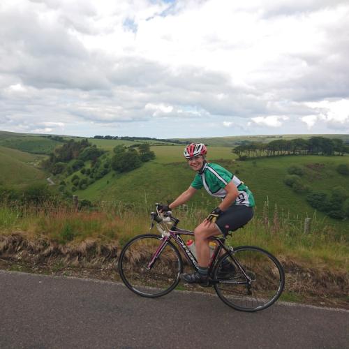 A woman is cycling on an Orbea road bike. She is wearing a green and white cycling jersey, black shorts, a red and white helmet and black and yellow fingerless gloves. She is smiling at the camera. In the background is some pretty impressive countryside