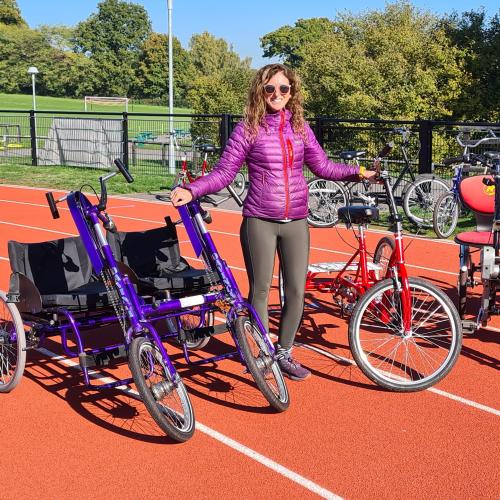 A woman is standing on a racetrack. She is wearing black leggings and a violet padded jacket, along with sunglasses. She has long, wavy, dark blonde hair. She is surrounded by various non-standard cycles, showing the different options for different needs