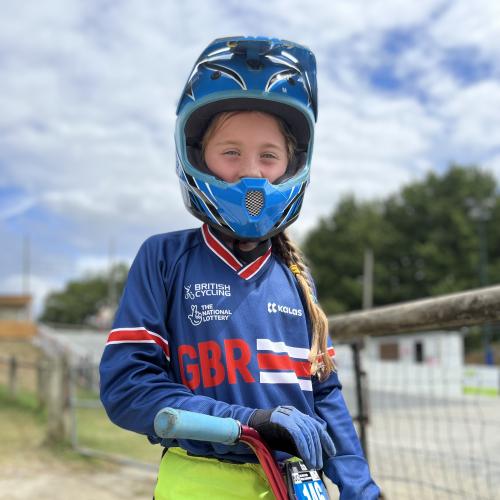 Amelie stands with her BMX bike. She is wearing a BMX helmet, gloves, blue British Cycling jersey and green trousers. 