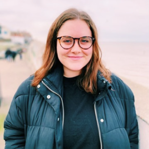 Amy stands with the beach behind her. She is wearing glasses and a dark jacket. 