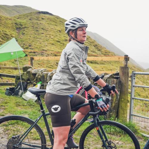 Charlotte stands side-on to the camera with her bike in a mountainous landscape. She is wearing a white helmet, grey jacket and brown shorts. 