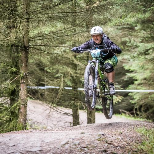 Emma is smiling as her bike does a jump on a mountain biking trail. She is wearing a helmet, dark coloured top and green leggings.