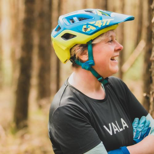 Emma smiles with her arms folded. She is wearing a blue and yellow helmet, black top and blue gloves. There are trees in the background.