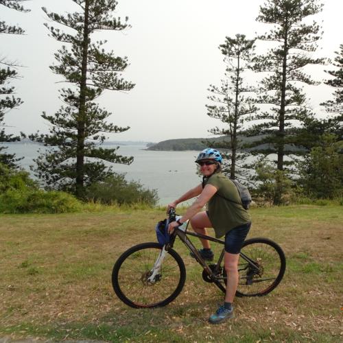 Helen smiling on her bike. She wears a blue helmet, green top and dark shorts. There is a lake behind her.