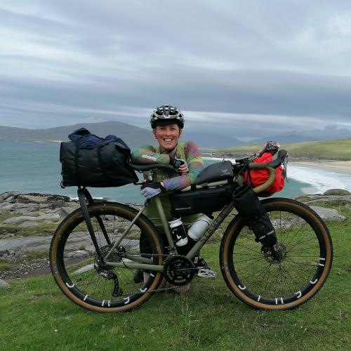 Iona kneels behind her green bike which is set up with bikepacking bags. She wears a helmet, green jersey, gloves and black shorts. A beach is in the background. 