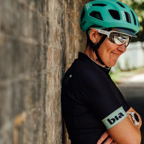 Jenni stands leaning against a brick wall with her arms folded. She wears a green helmet, sunglasses and black jersey. 