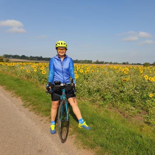 Marina stands next to a sunflower field with her blue bike. She wears sunglasses, a yellow helmet, blue jacket and black shorts. 