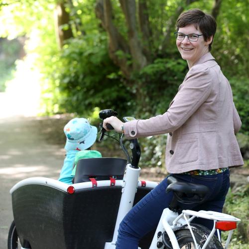 Saskia smiles at the camera as she stands with her cargo bike, transporting her children. She is wearing glasses, a beige jacket and jeans. 