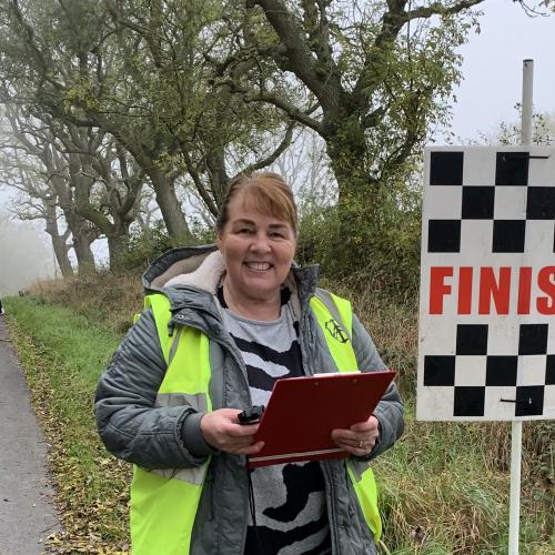 Sharon smiles to camera, holding a clipboard and stopwatch. She is standing at the side of the road at the finish line. She is wearing a grey winter coat and hi-vis vest.