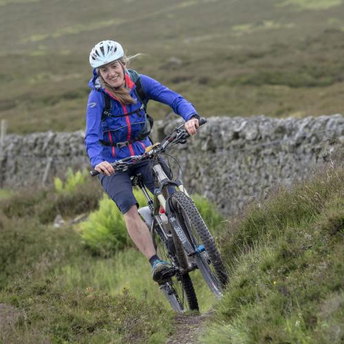 Sophie mountain biking on the Great North Trail. She is wearing a white helmet, blue jacket and grey shorts. 